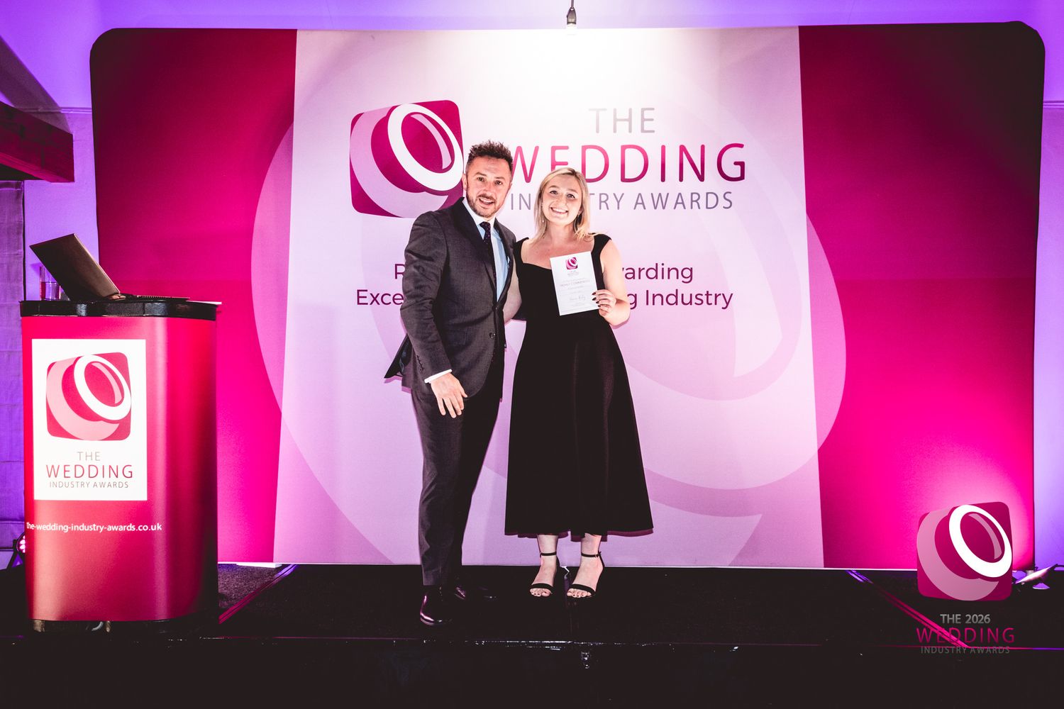 Event stage setup with pink lighting and corporate branding displays at a wedding industry conference.