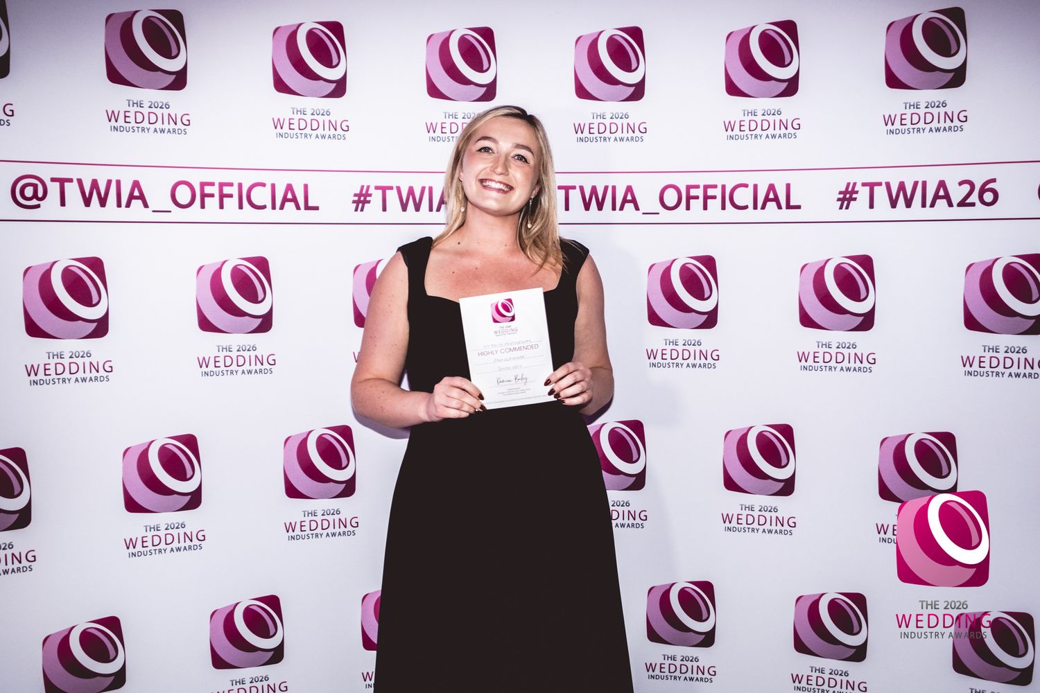 A person poses on a red carpet against a white backdrop with repeating pink logos at an awards event.
