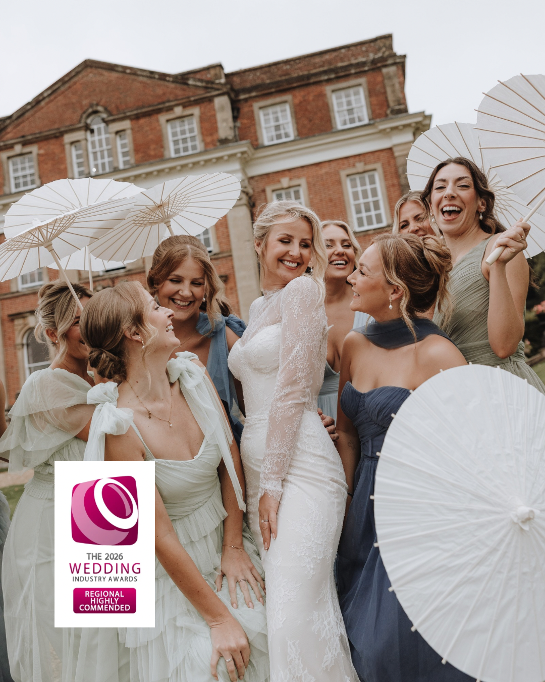 A joyful group of bridesmaids in sage green dresses share a candid moment with the bride outside a brick building.