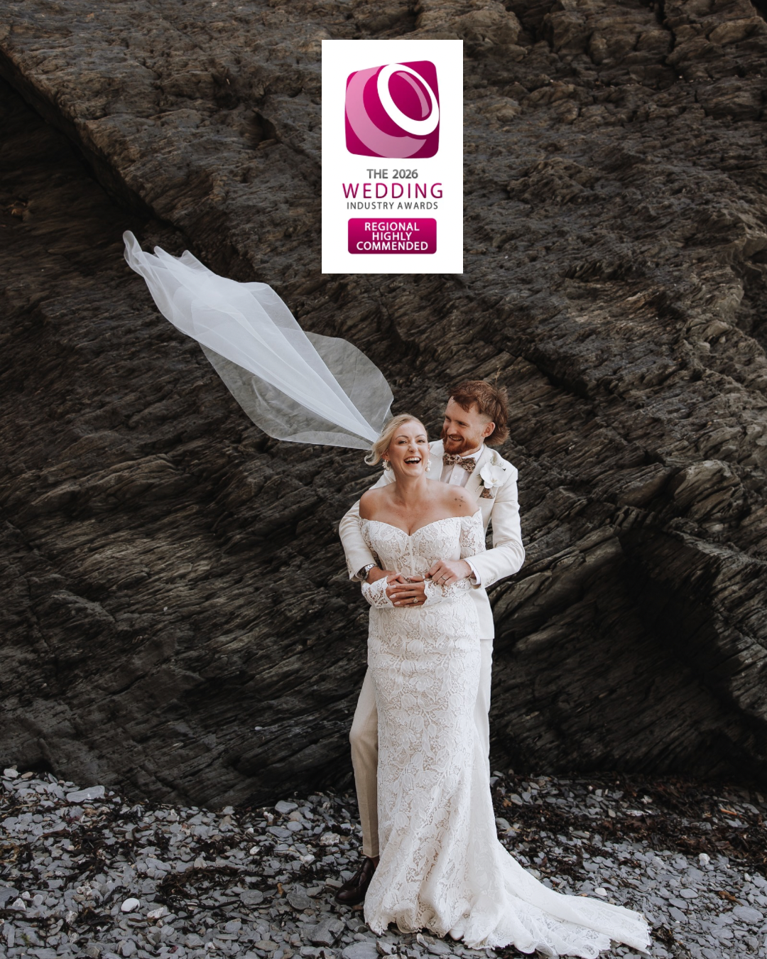 A couple in wedding attire embraces on dark rocky terrain as a white veil flows dramatically in the wind.