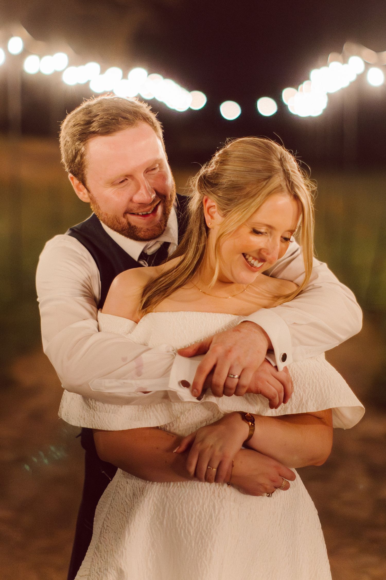A romantic outdoor wedding scene with string lights overhead, featuring a couple on a dirt path at night.