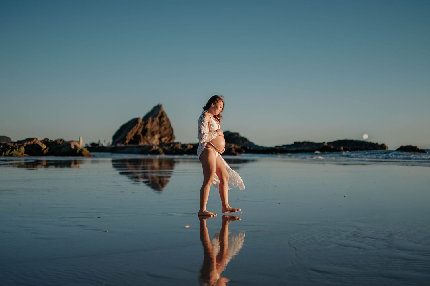 A silhouette of a person walks along a reflective beach at sunset with coastal rock formations in the background.