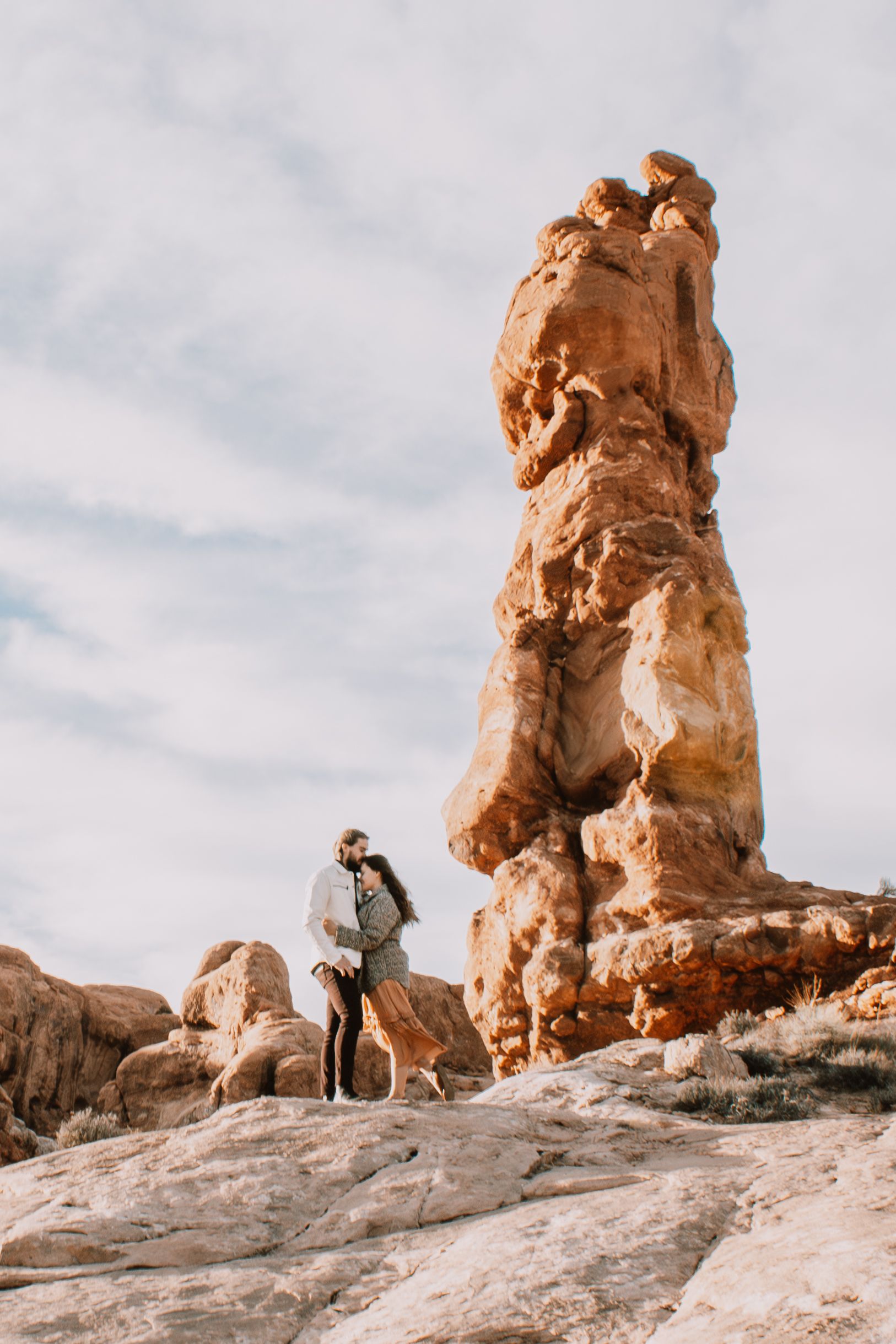 Couple kissing beside a towering red rock formation under a cloudy sky in the desert.