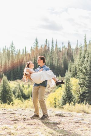 Man carrying woman in his arms in a sunny mountain meadow surrounded by evergreen and autumn trees.