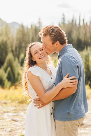 A couple embraces outdoors, woman in white dress smiling as man kisses her cheek, with pine trees in background.