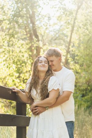 Man embracing woman from behind as she leans against a wooden fence in a bright, sunny forest.
