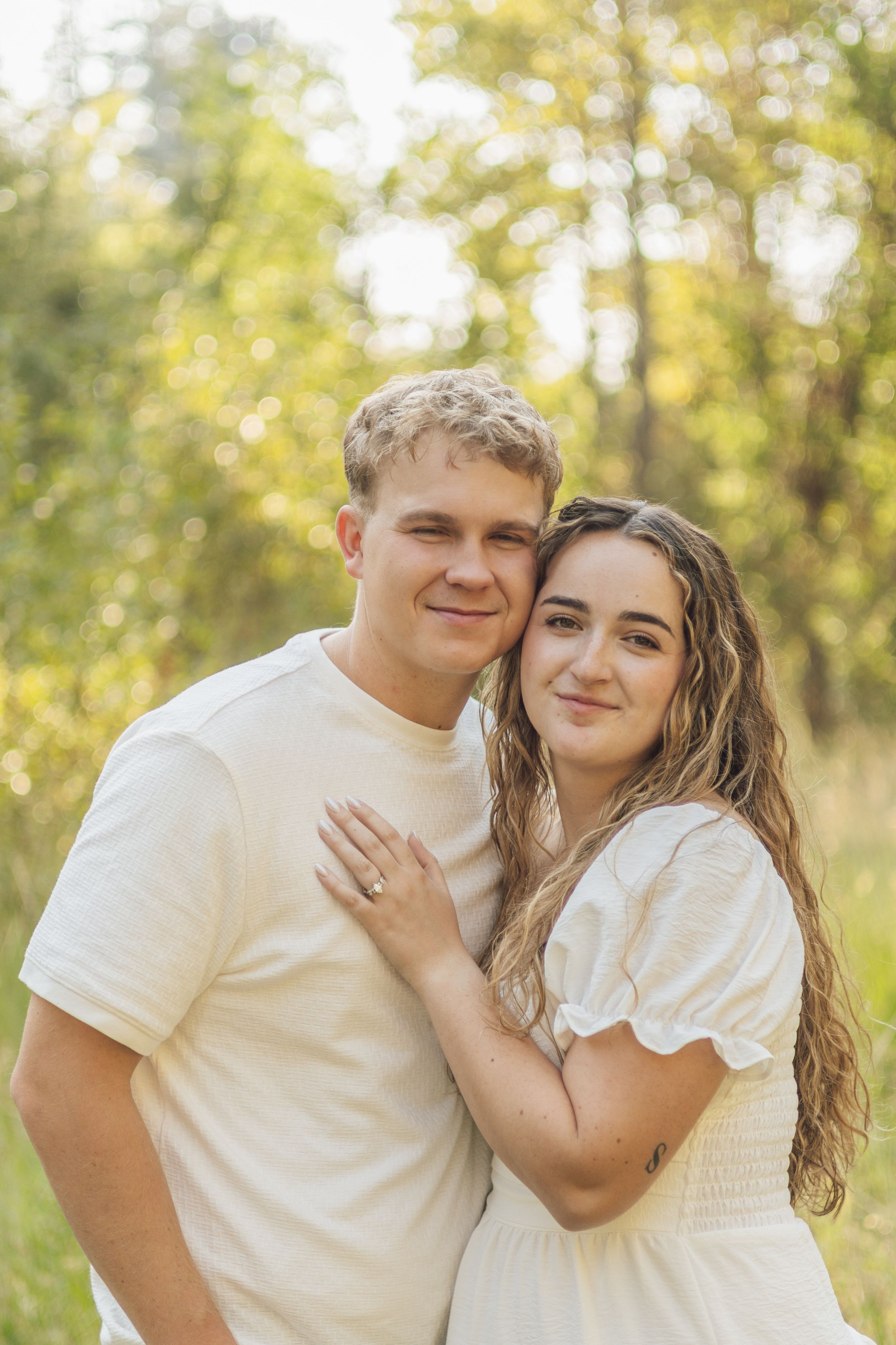 Smiling couple posing together in a sun-kissed outdoor setting with lush green trees behind them.