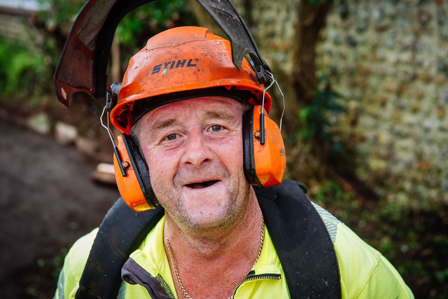 Construction worker wearing orange safety gear and ear protection smiles while working outdoors.