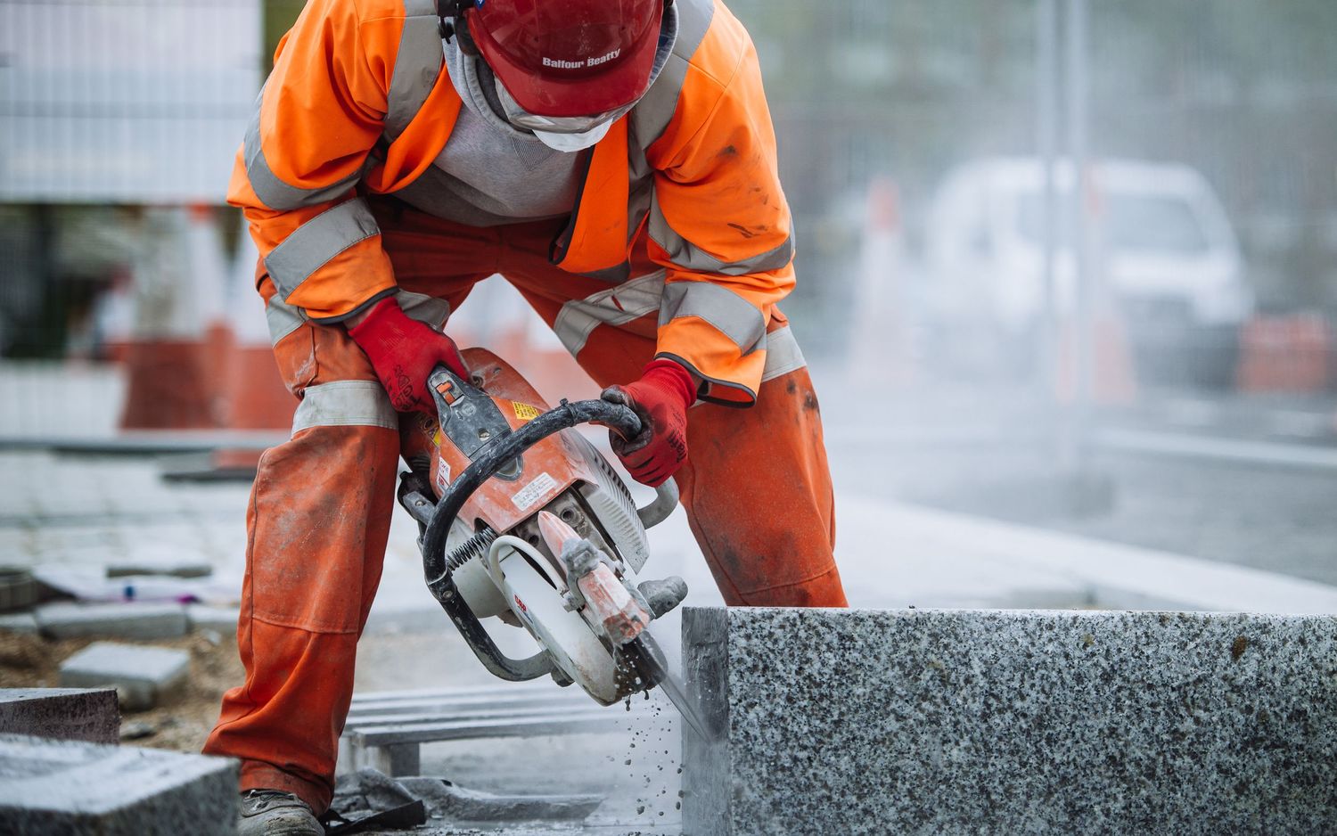 Worker in orange safety gear cutting concrete with power tools creating dust spray.