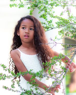 Young child in white dress poses playfully near green foliage in a bright outdoor setting.