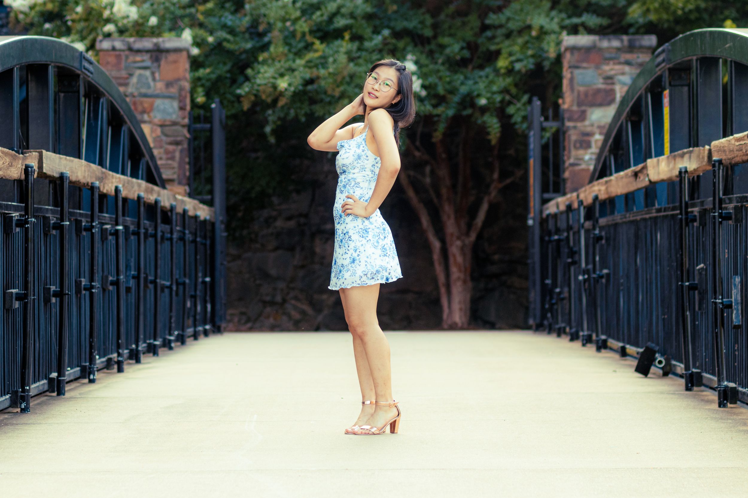 A person performs a graceful gymnastic pose on a bridge walkway surrounded by lush green trees.
