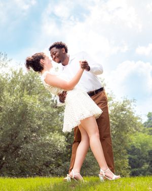 A couple in formal attire dips gracefully while dancing together on a grassy field under blue skies.
