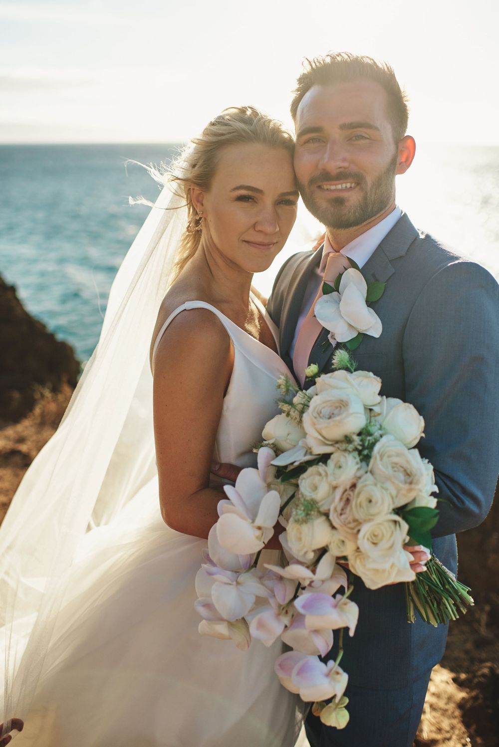 Coastal wedding portrait with elegant white bouquet against ocean backdrop at sunset.