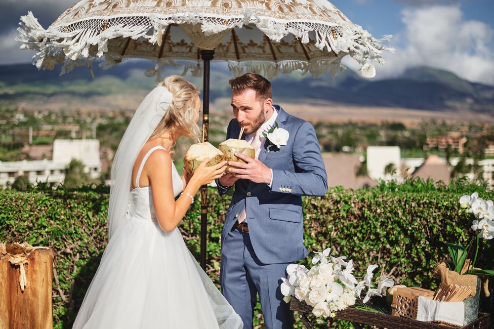 Newlyweds share a drink under a rustic umbrella at an outdoor vineyard wedding with mountains in the background.