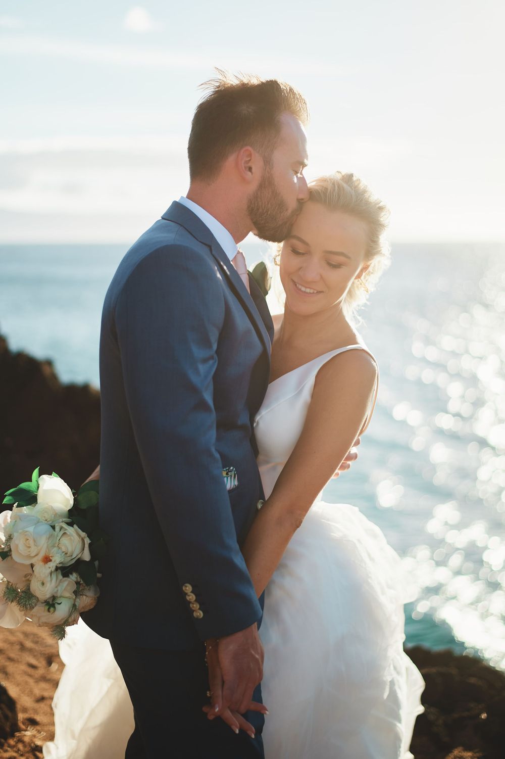 Intimate moment between newlyweds on oceanside cliff during golden hour.