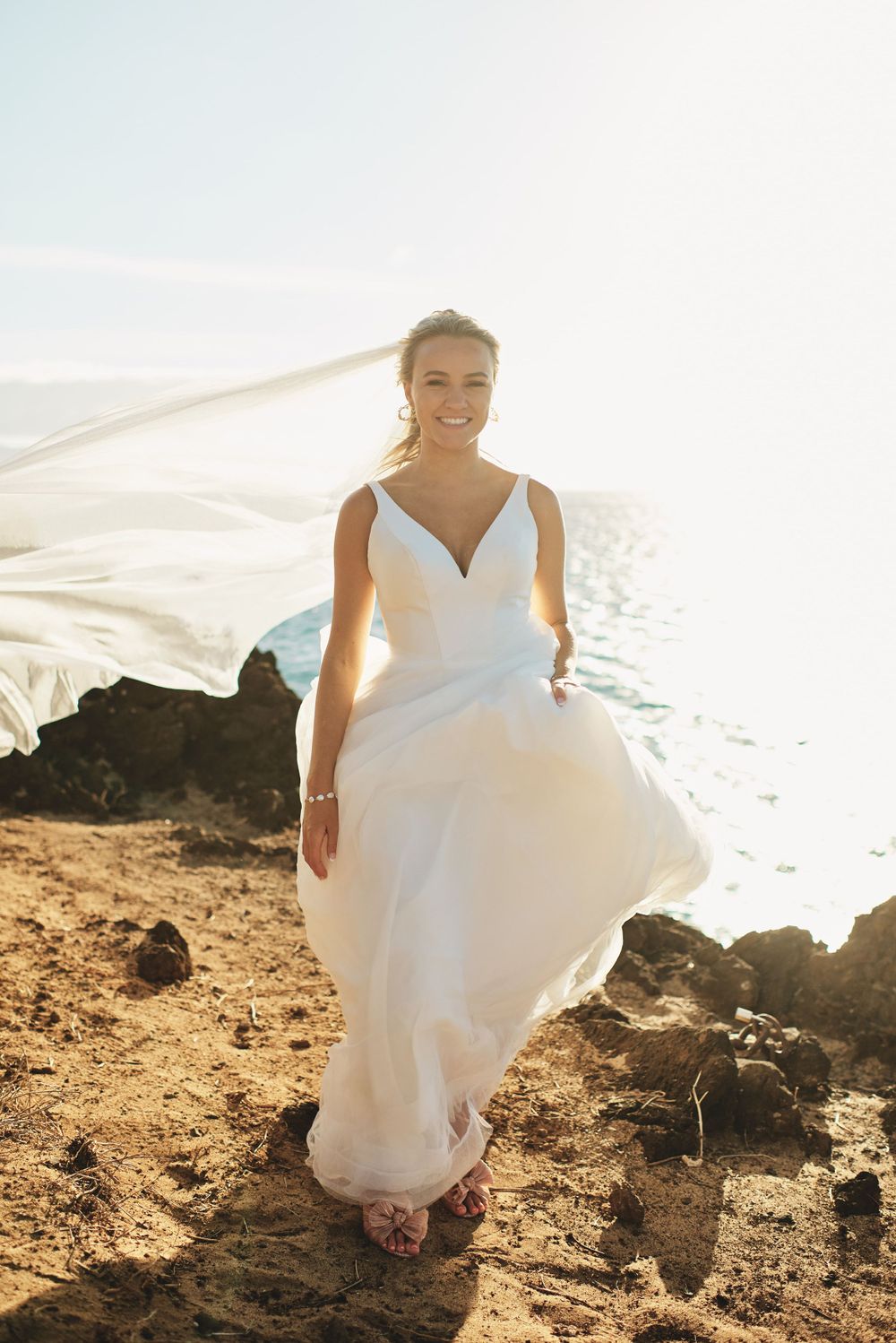 Bride in flowing white dress poses against sparkling ocean backdrop.