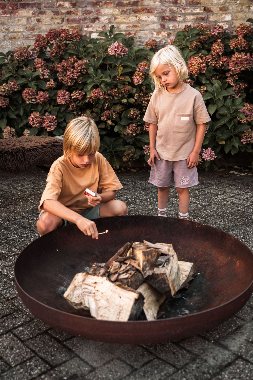 Children gathered around a rustic fire pit with flowering bushes in the background.