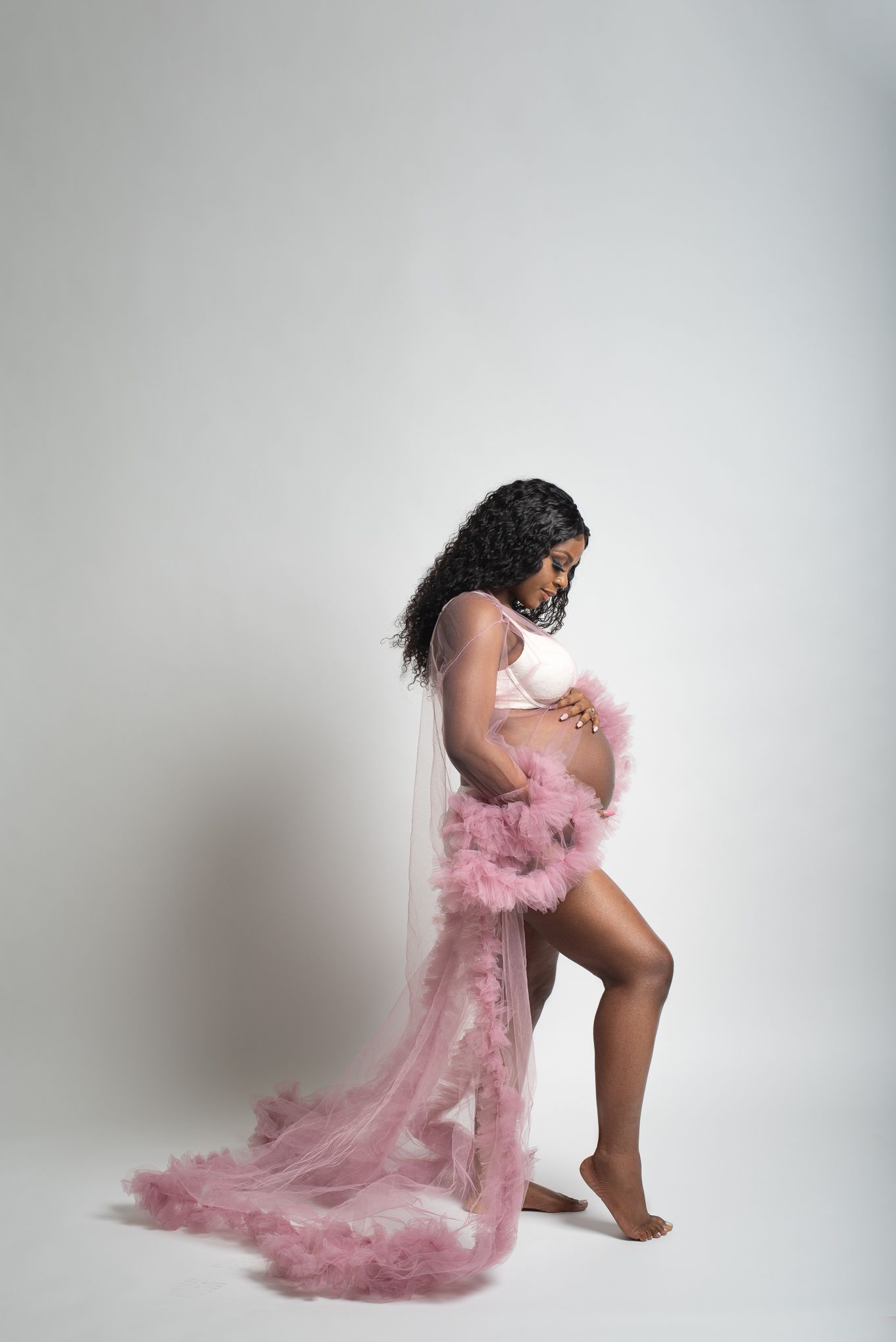 Elegant maternity portrait in white off-shoulder flowing dress against brown backdrop, showcasing a graceful pose.