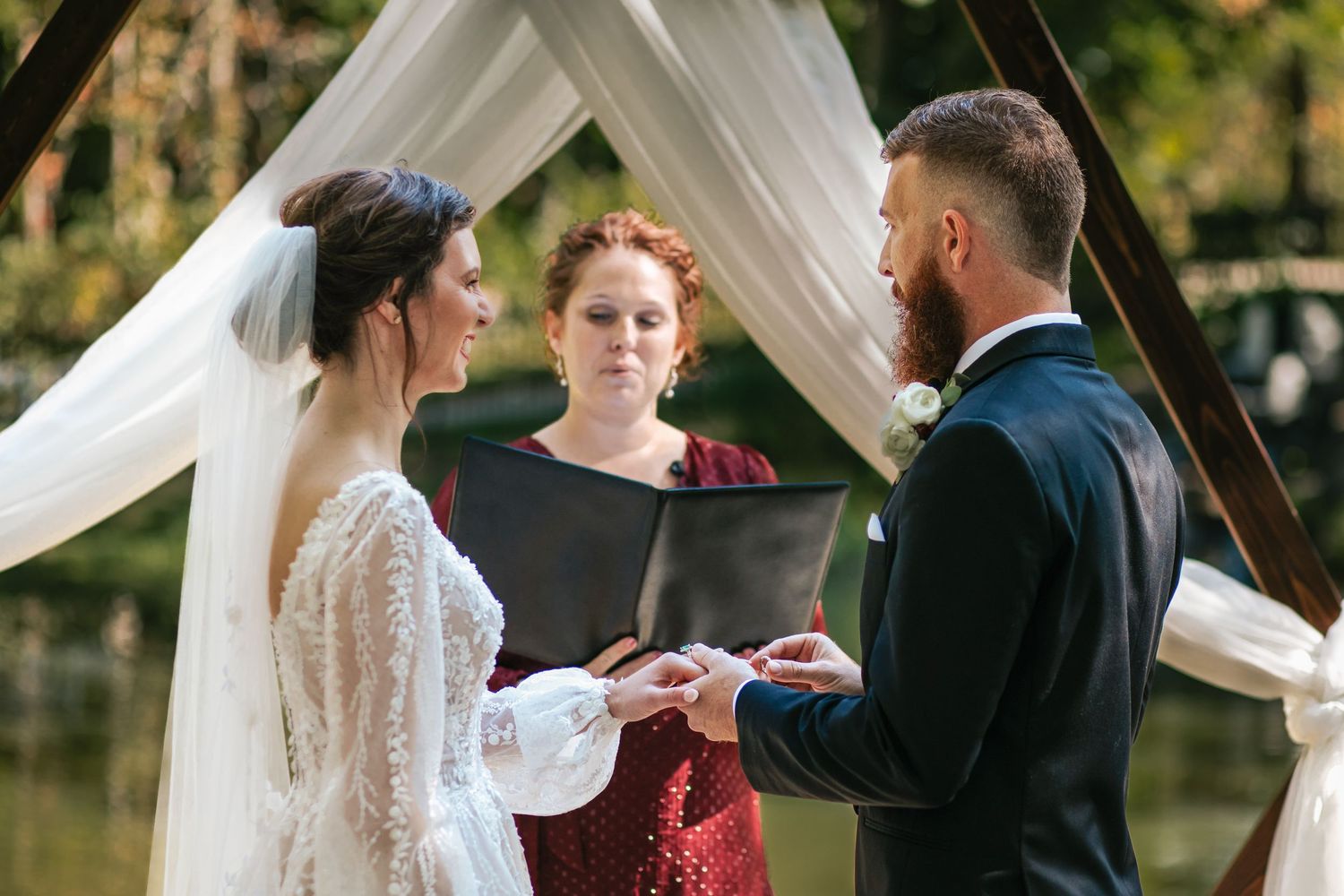 An outdoor wedding ceremony with white drapery and guests seated on white chairs in a wooded setting.