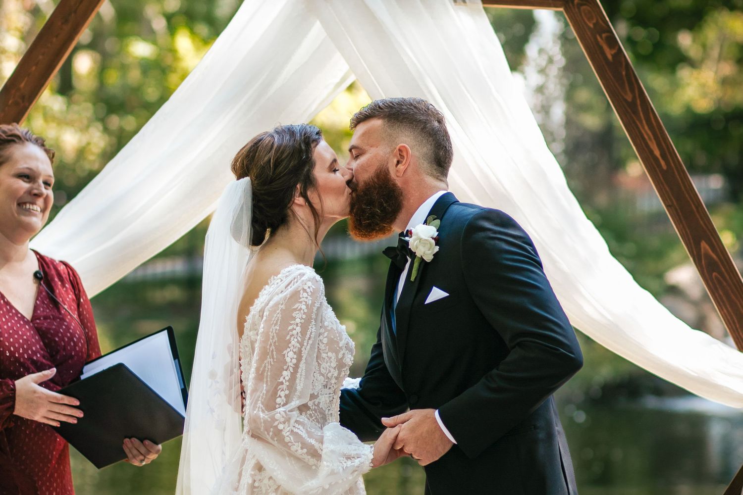 A wedding ceremony takes place outdoors under white fabric draping with autumn foliage in the background.