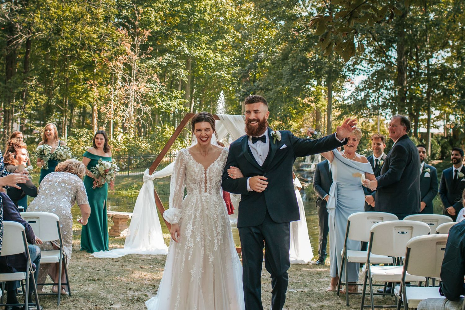 Wedding guests gather for an outdoor ceremony with white chairs and autumn trees in the background.