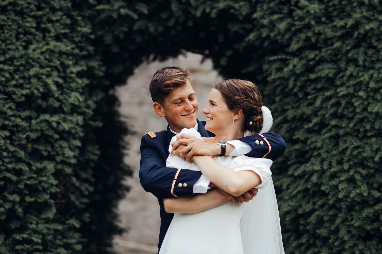 Military groom in uniform embracing smiling bride in white dress before lush green hedge arch backdrop.