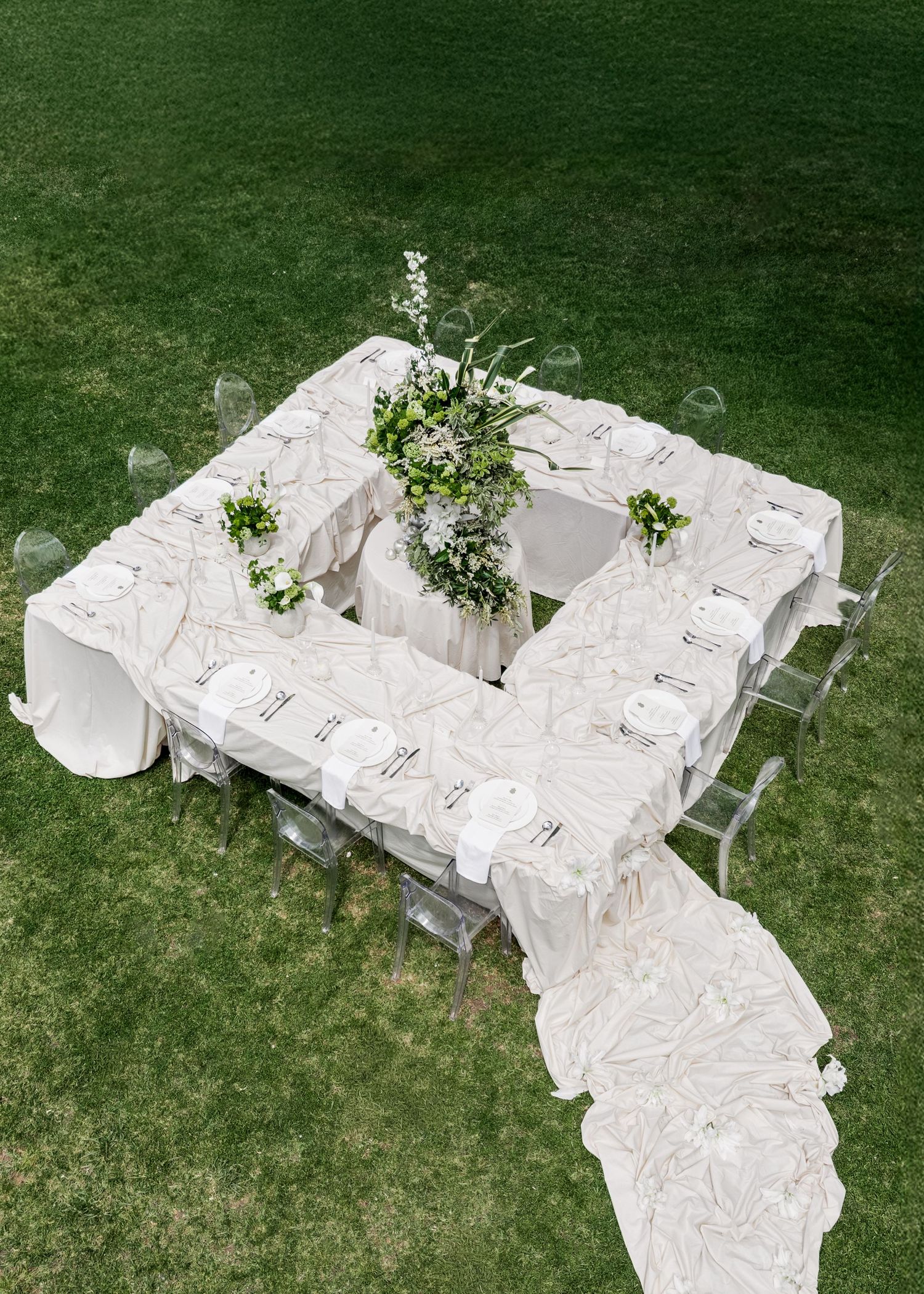 Elegant white tablecloth-draped U-shaped dining table with floral centerpieces set up on green grass for a wedding Al Ru Farm in Adelaide.