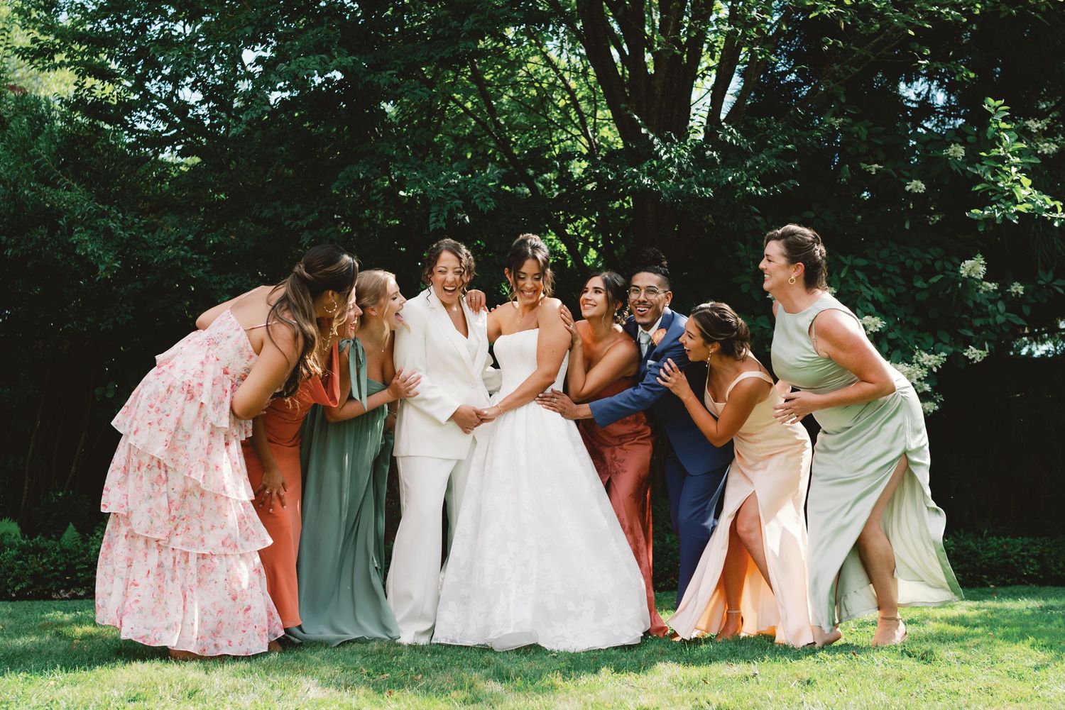 Group of bridesmaids in flowing dresses pose together on grassy lawn during outdoor wedding celebration.