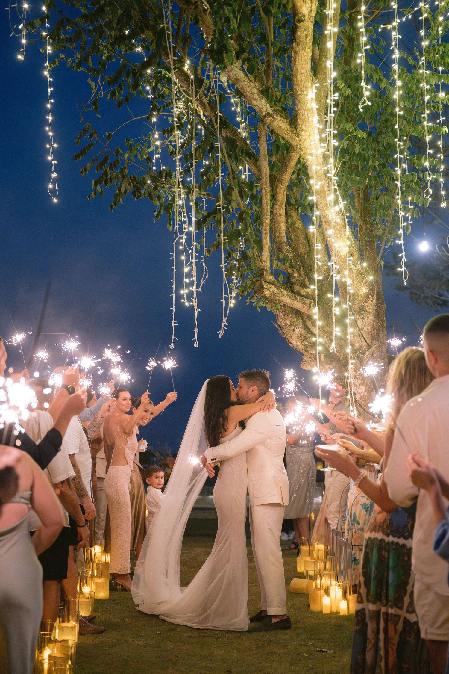 Wedding guests in white attire dance and celebrate under trees decorated with hanging string lights at dusk.