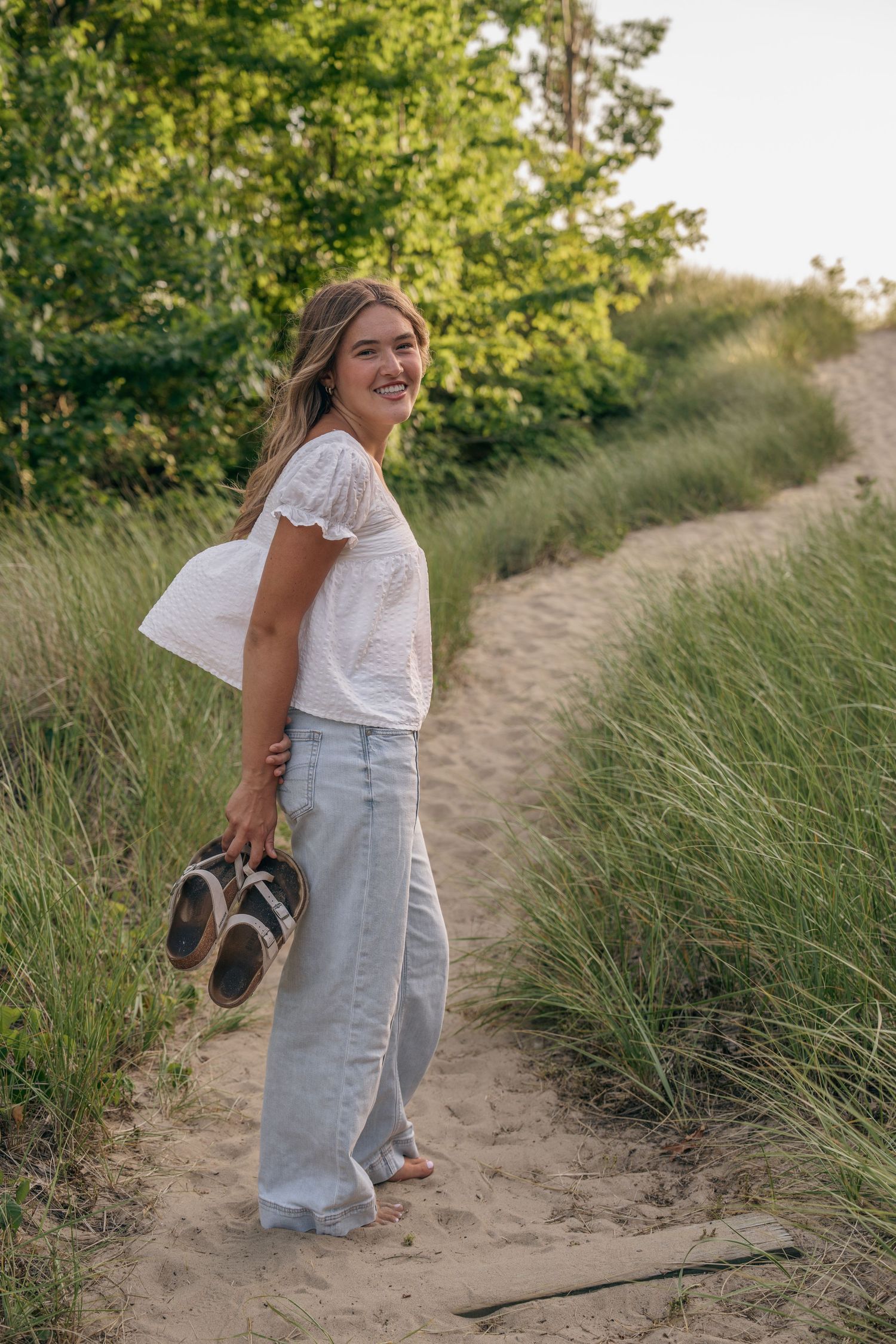 Smiling woman in white top and light jeans holding sandals on a sandy dune path surrounded by green grass.