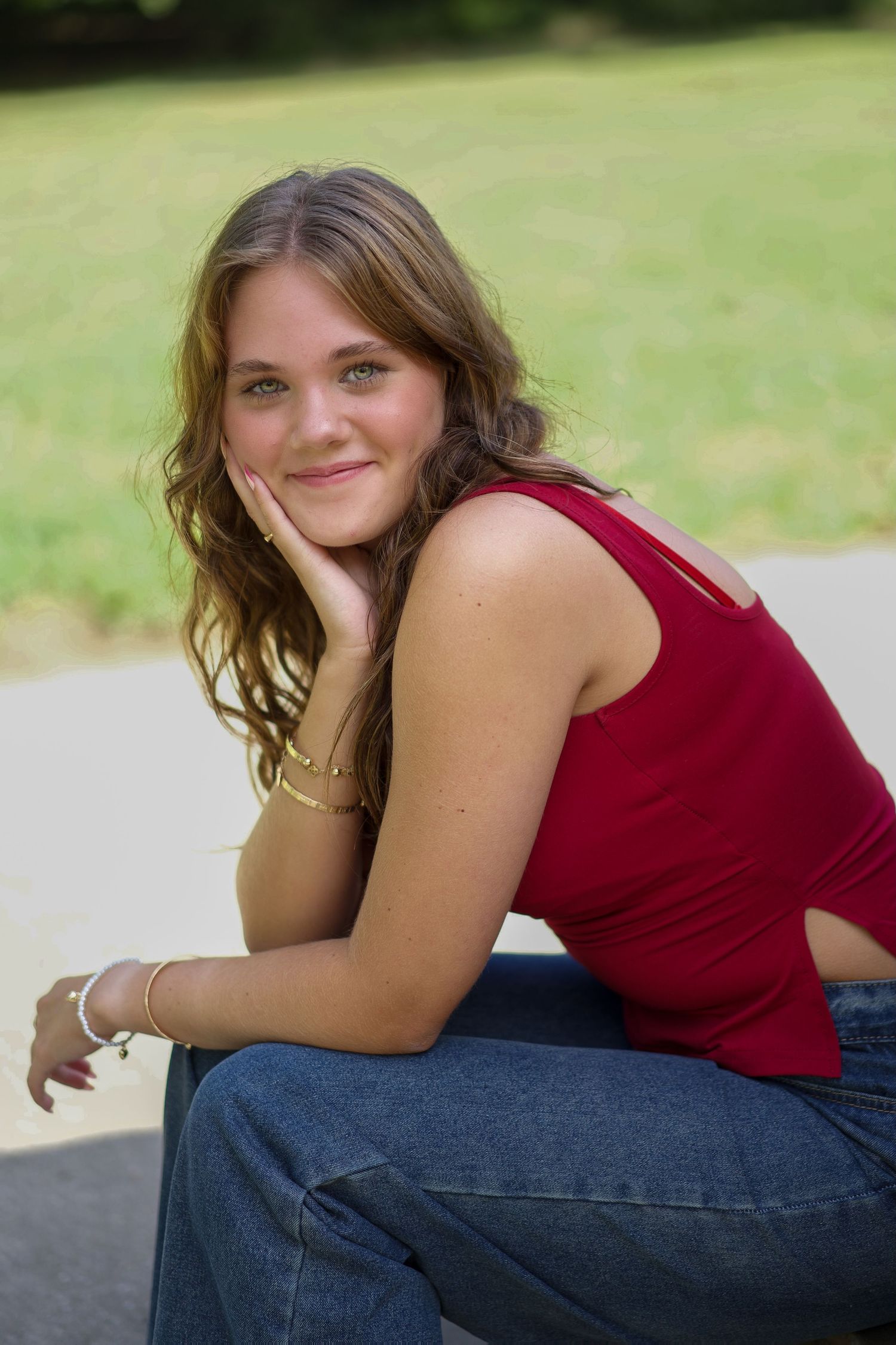 Someone in a red sleeveless top and jeans poses casually outdoors on a sunny day with a green background.