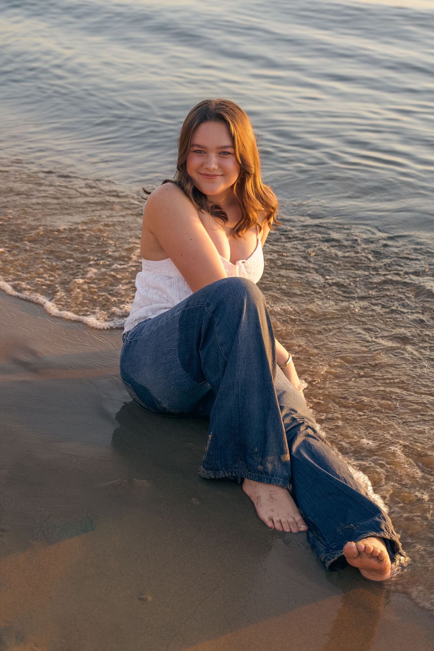 Person in white top and blue jeans sits at water's edge during sunset at the beach.
