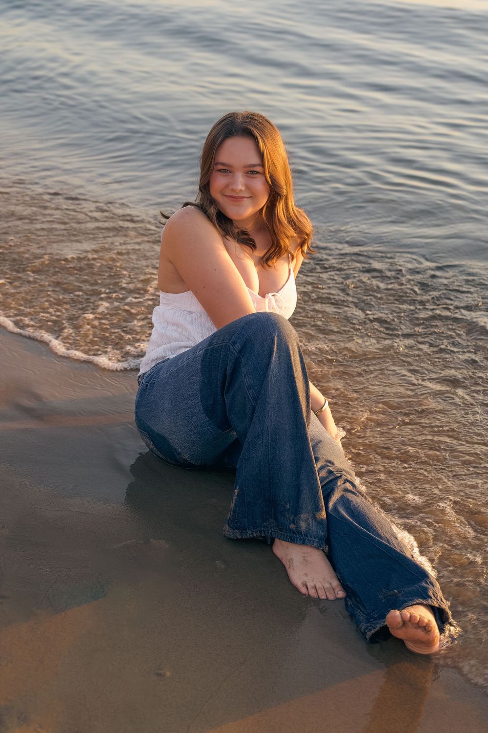Person in white top and blue jeans sits at water's edge during sunset at the beach.