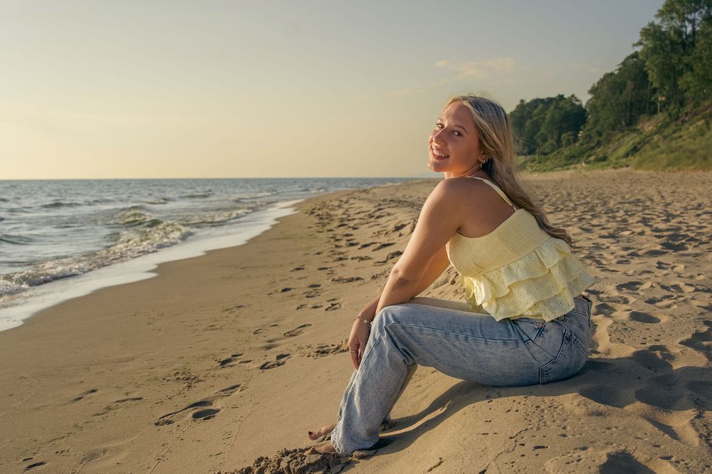 Person in yellow top and jeans sits on sandy beach at sunset enjoying peaceful ocean view.