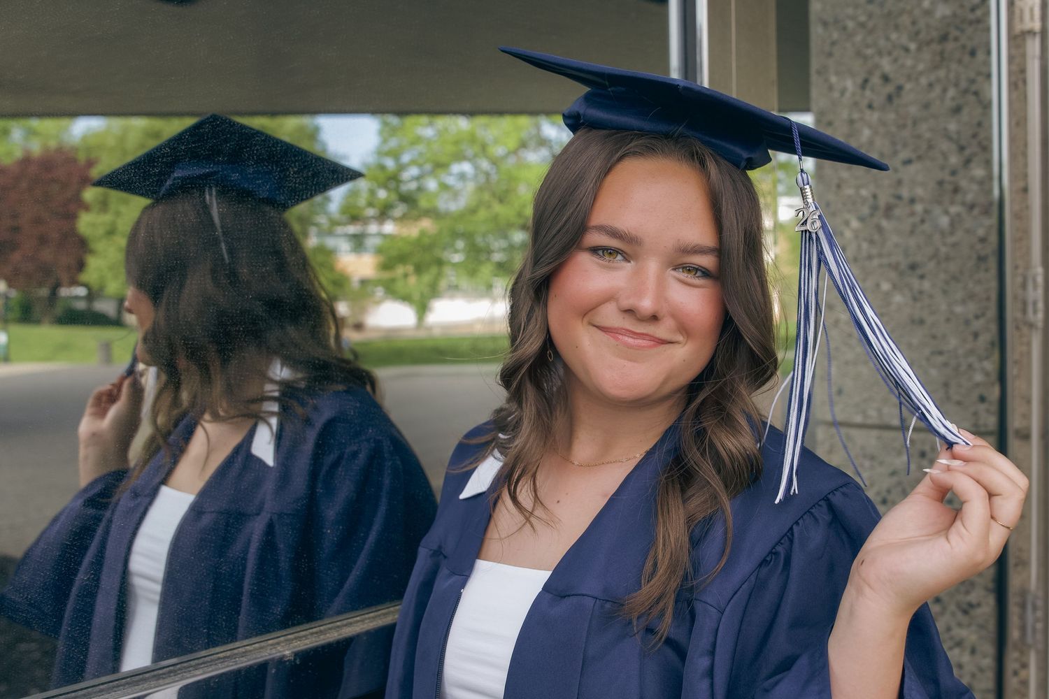 Smiling young woman in blue graduation cap and gown posing near reflective glass surface.
