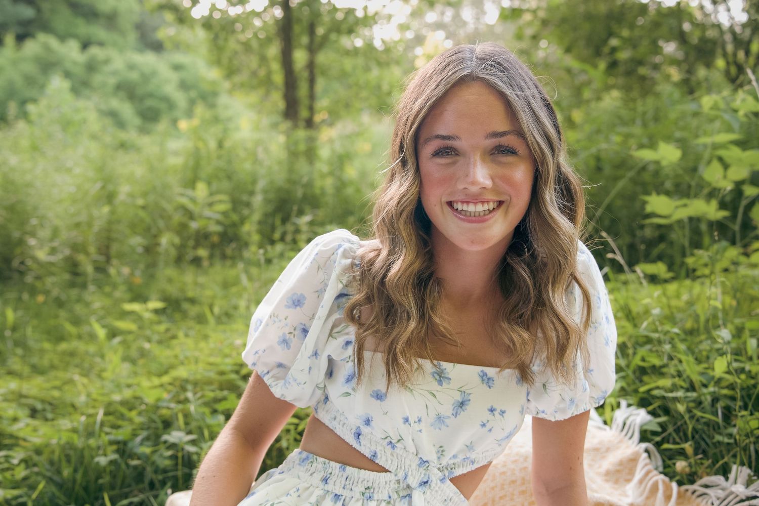 A person in a floral dress poses outdoors in a garden setting with lush green foliage in the background.