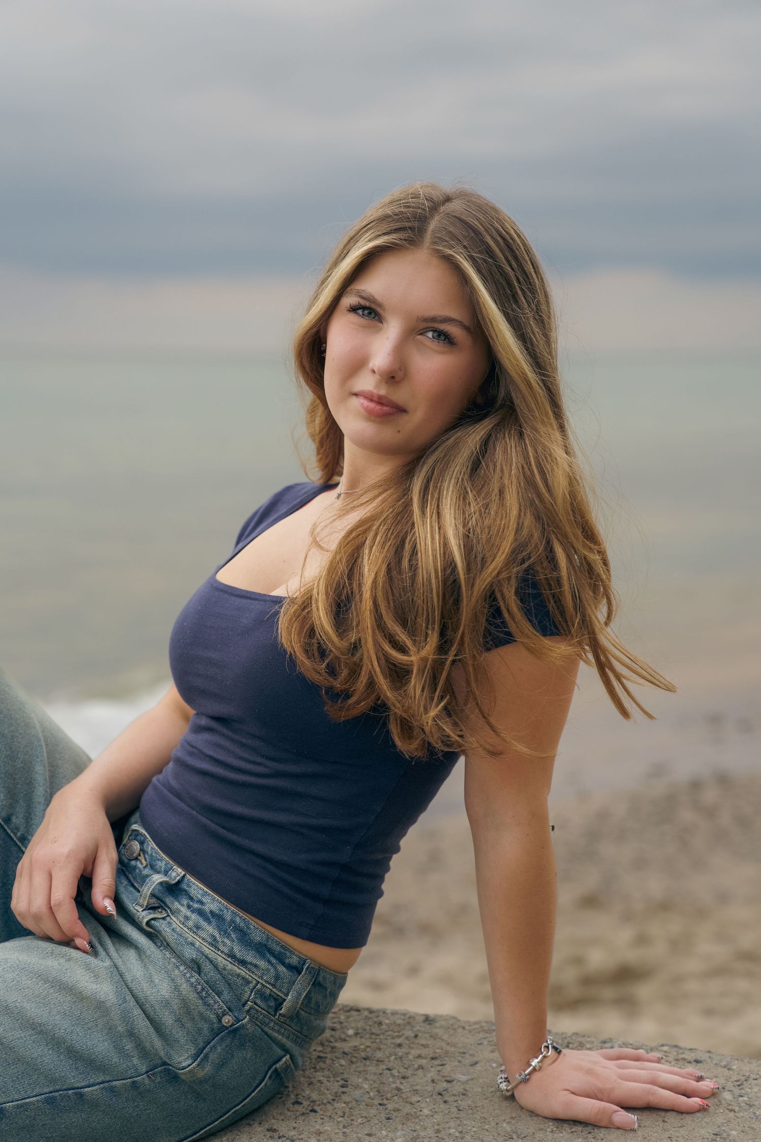A person in a navy tank top and jeans sits casually on a beach with water and cloudy skies in the background.
