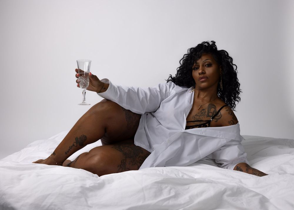 A person in a white shirt lounges on white bedding while holding a glass in a sensual boudoir photo.