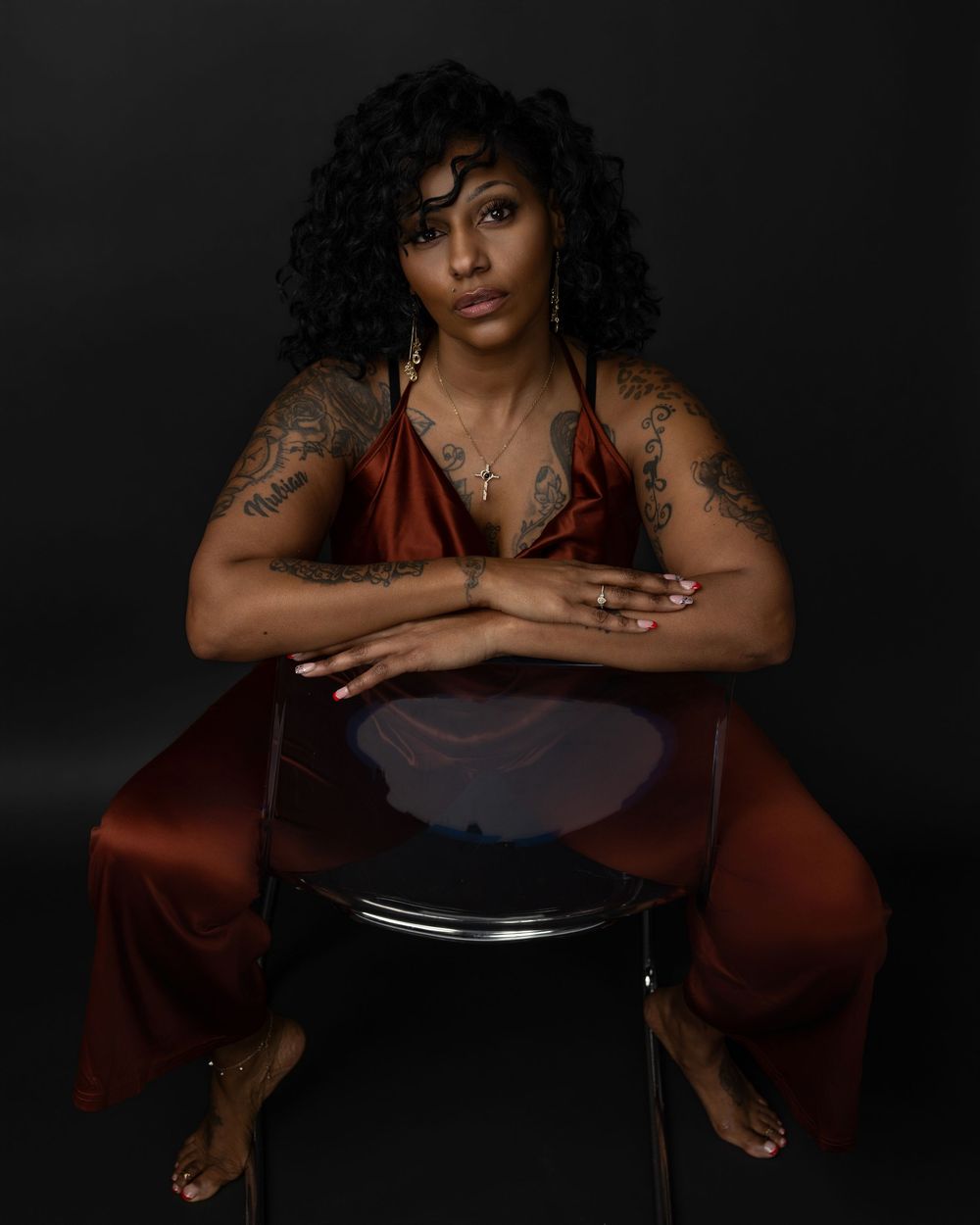 Artistic portrait series showing figure in dark setting seated on chair with curly hair and tattoos against black background.