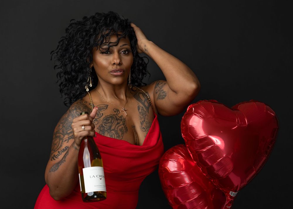 A person in a glamorous red dress poses against a dark background with heart-shaped balloons and dramatic lighting.