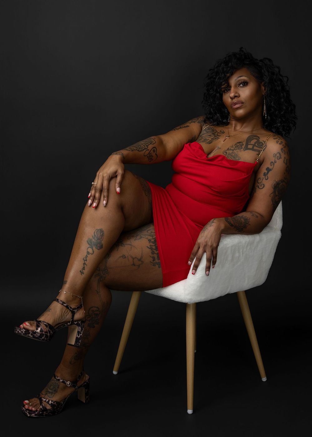 A model in a red dress poses elegantly on a white chair against a dark studio backdrop.