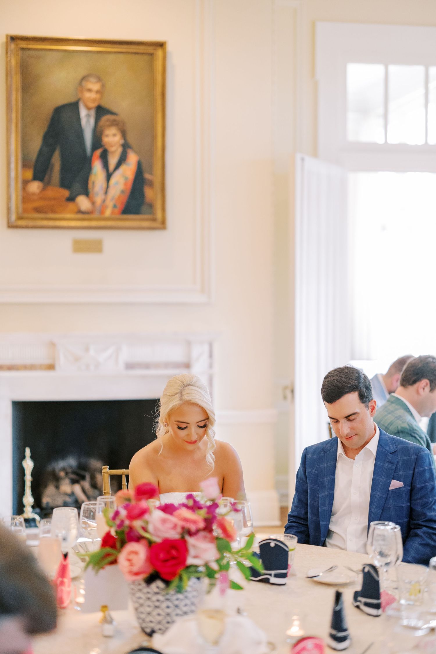 Elegant wedding reception dinner table with pink floral centerpieces and guests seated in a bright room with framed portraits.