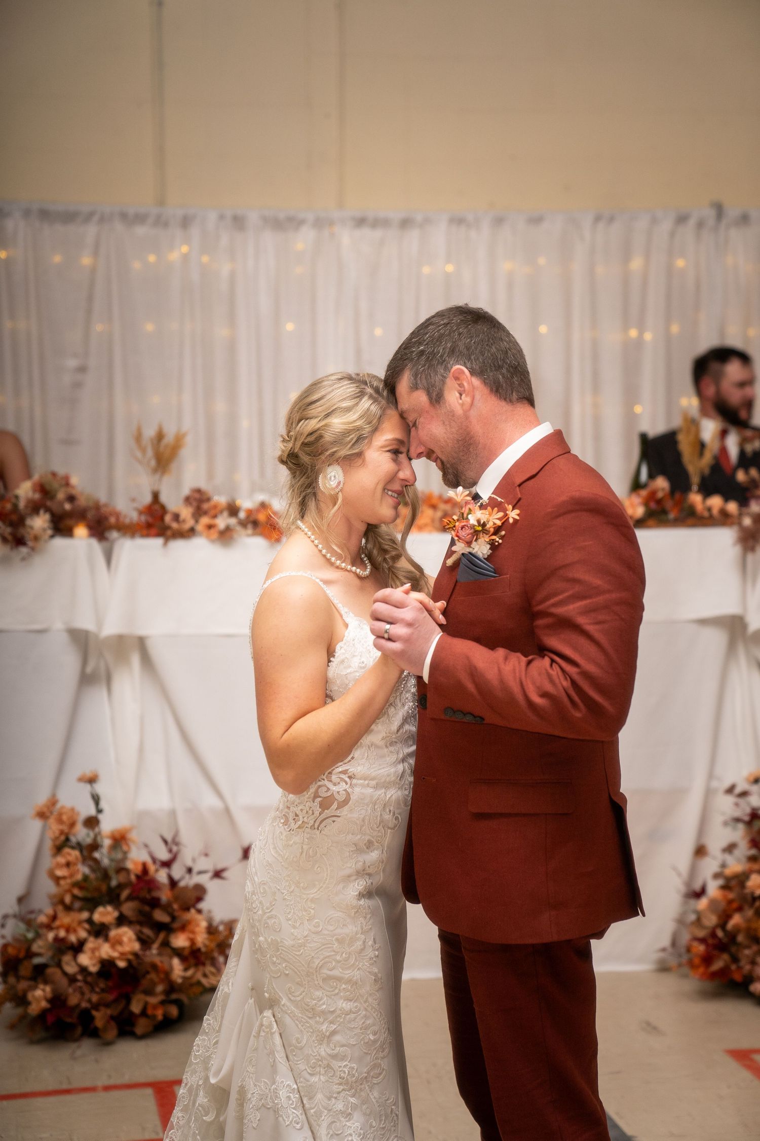A couple shares their first dance at a wedding reception decorated with autumn-colored floral arrangements.
