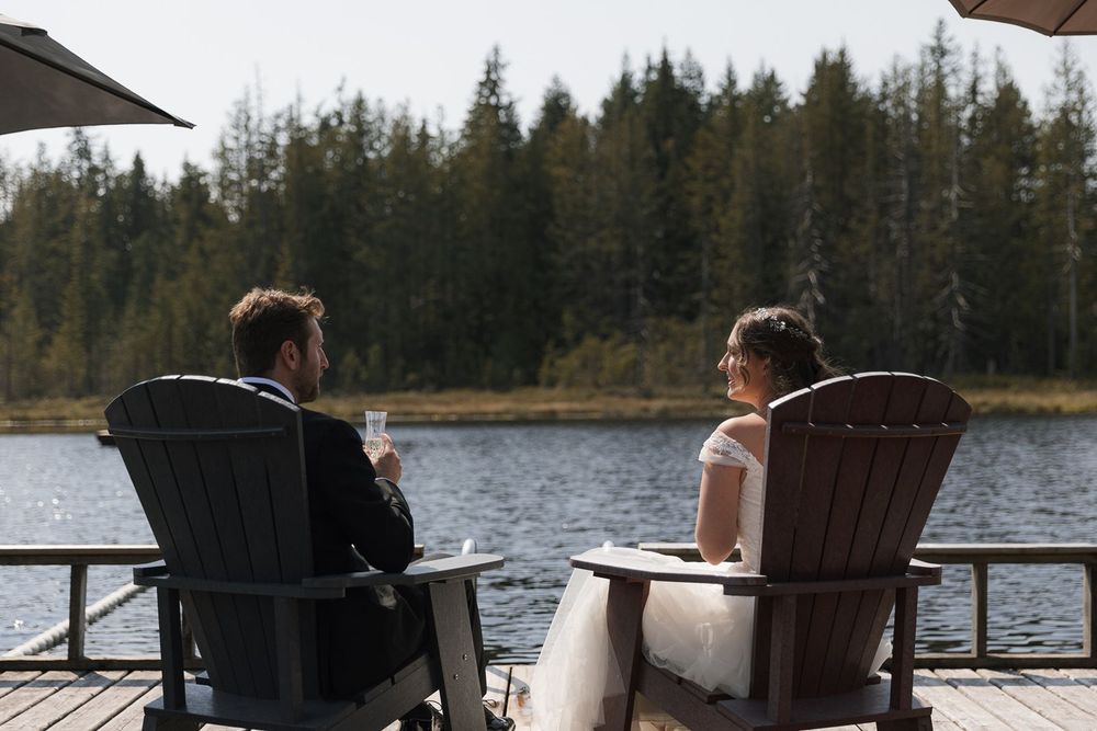 Bride and groom sitting in Adirondack chairs on a lakeside dock enjoying drinks at their wedding.