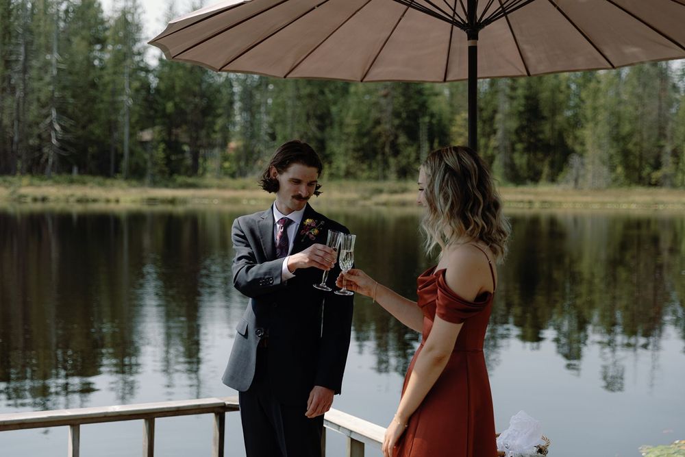 Bride and groom toasting with champagne glasses on a lakeside deck under a patio umbrella.