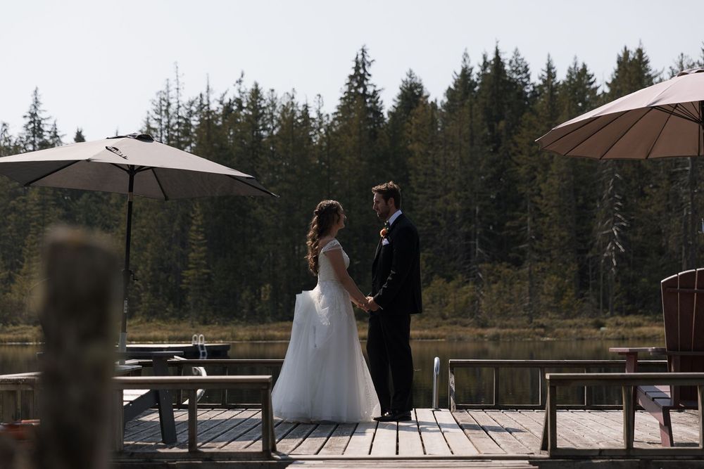 Bride and groom holding hands on a wooden dock surrounded by forest and a serene lake.