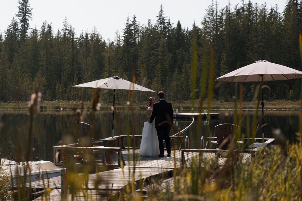 Bride and groom standing together on a lakeside dock with patio umbrellas and tall grass.