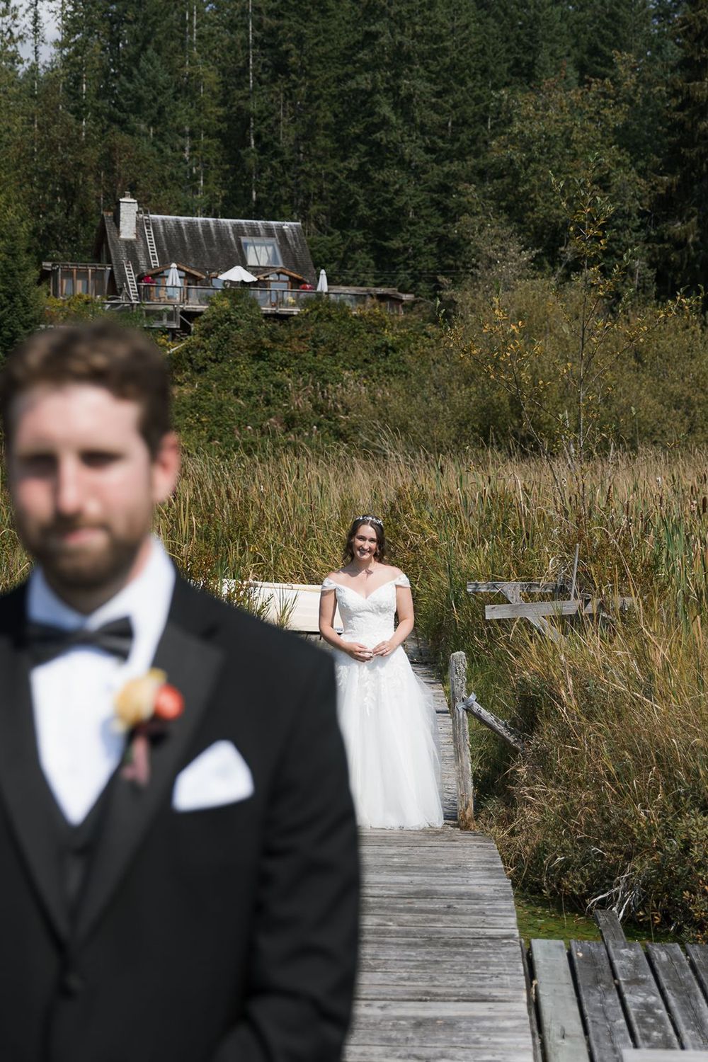 Bride in white gown smiling on a wooden boardwalk with groom in foreground at outdoor wedding.