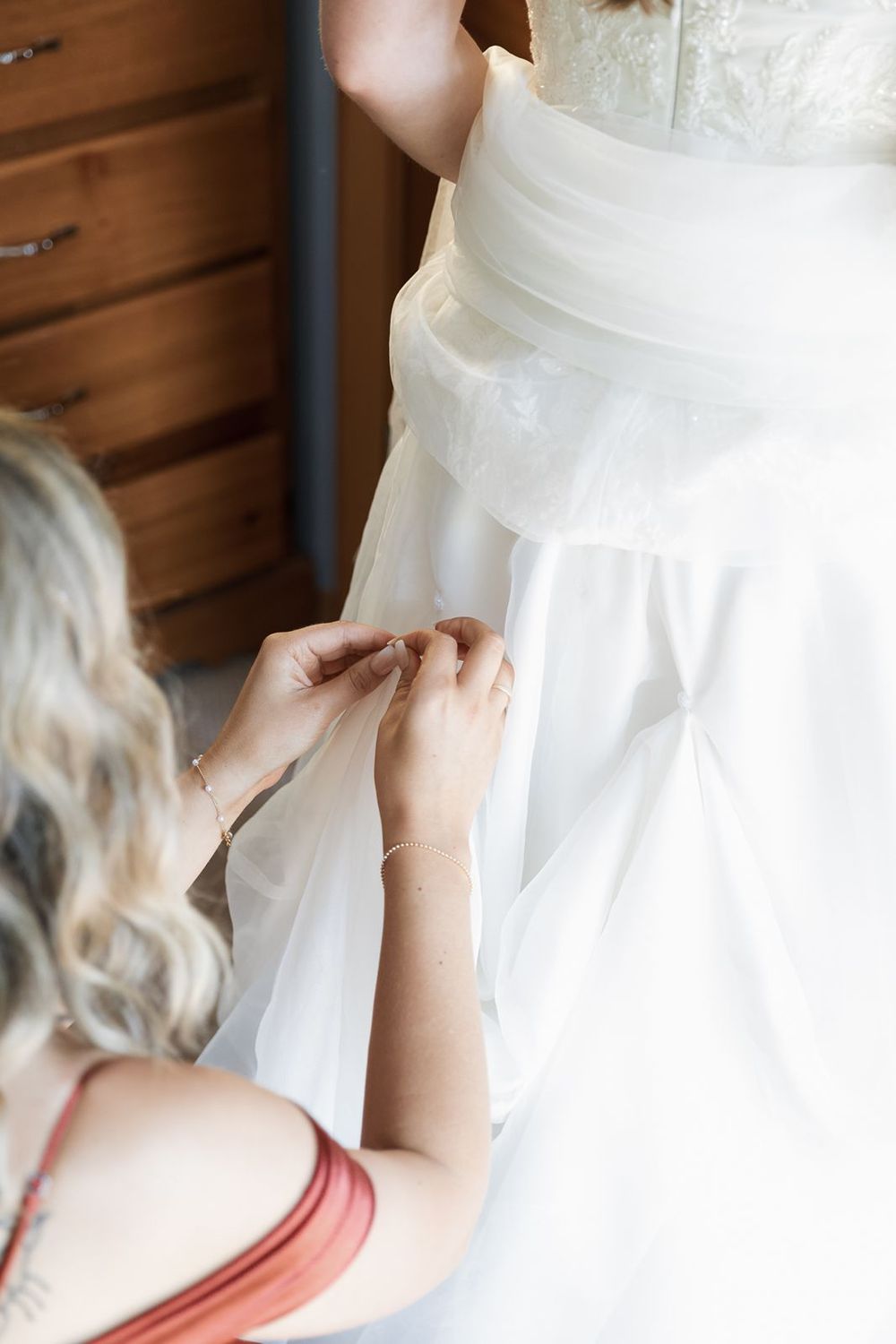 Bridesmaid in red dress fastening buttons on the back of a white wedding gown.