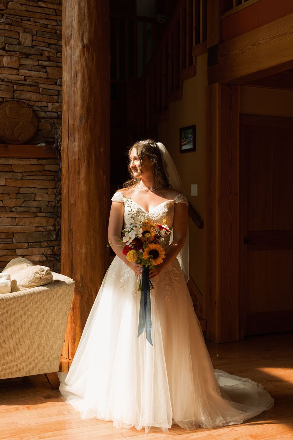 Radiant bride in lace off-shoulder gown holds colorful sunflower bouquet while posing in rustic log cabin interior.